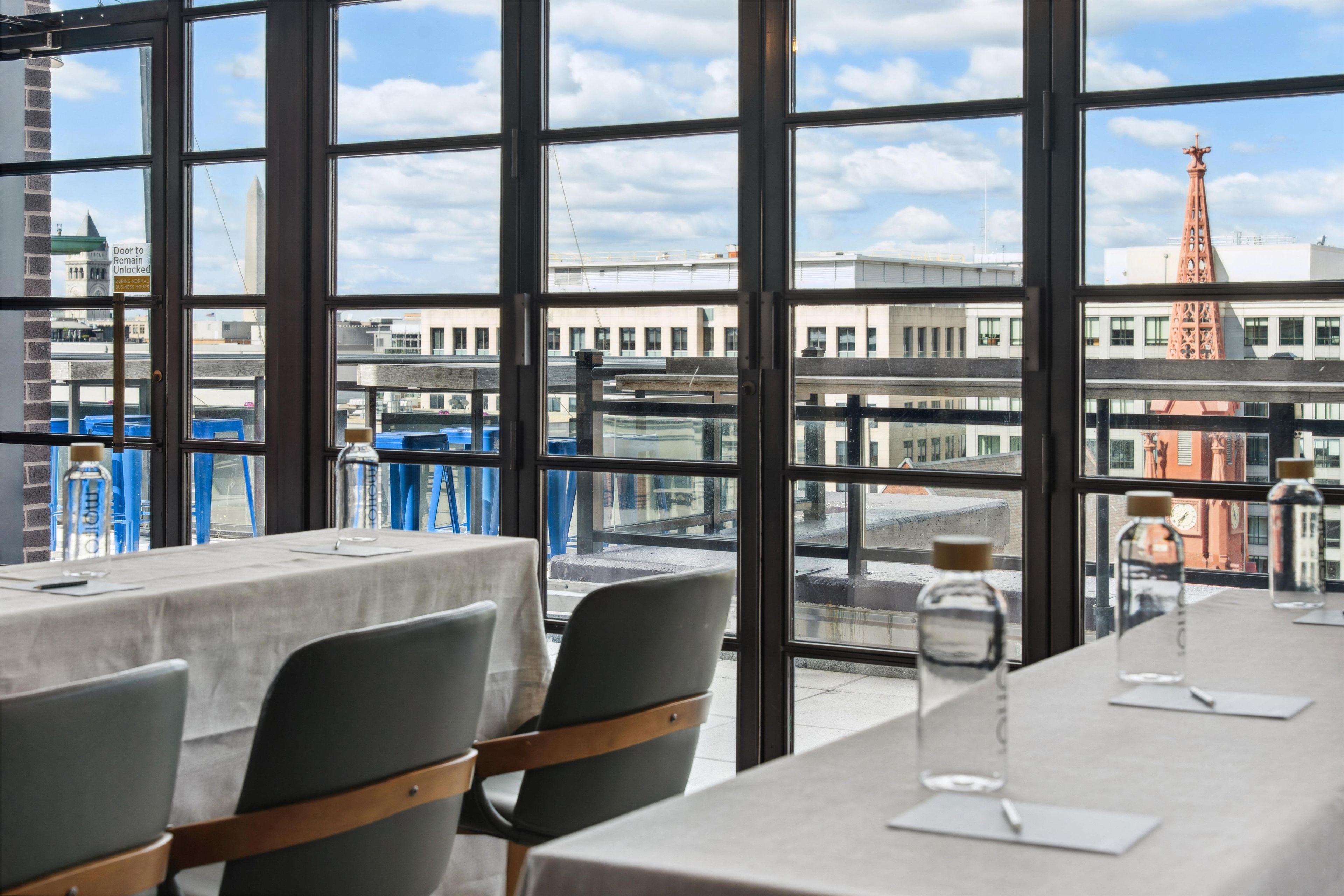 Ein Restaurant mit gedeckten Tischen, großen Fenstern mit Blick auf die Stadt und wolkenlosem Himmel.