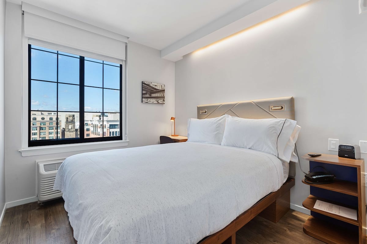 Ein modernes Schlafzimmer mit großem Fenster, einem Bett, einem Nachttisch und einem wandmontierten Regal.