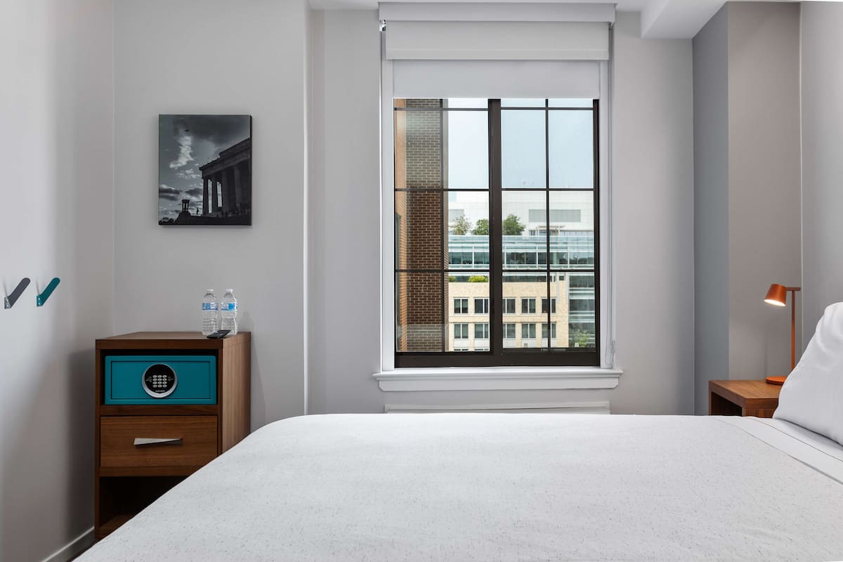 Ein Hotelzimmer mit einem großen Bett, einem Nachttisch mit Lampe, einem Fenster mit Blick auf Gebäude und einem Gemälde an der Wand.