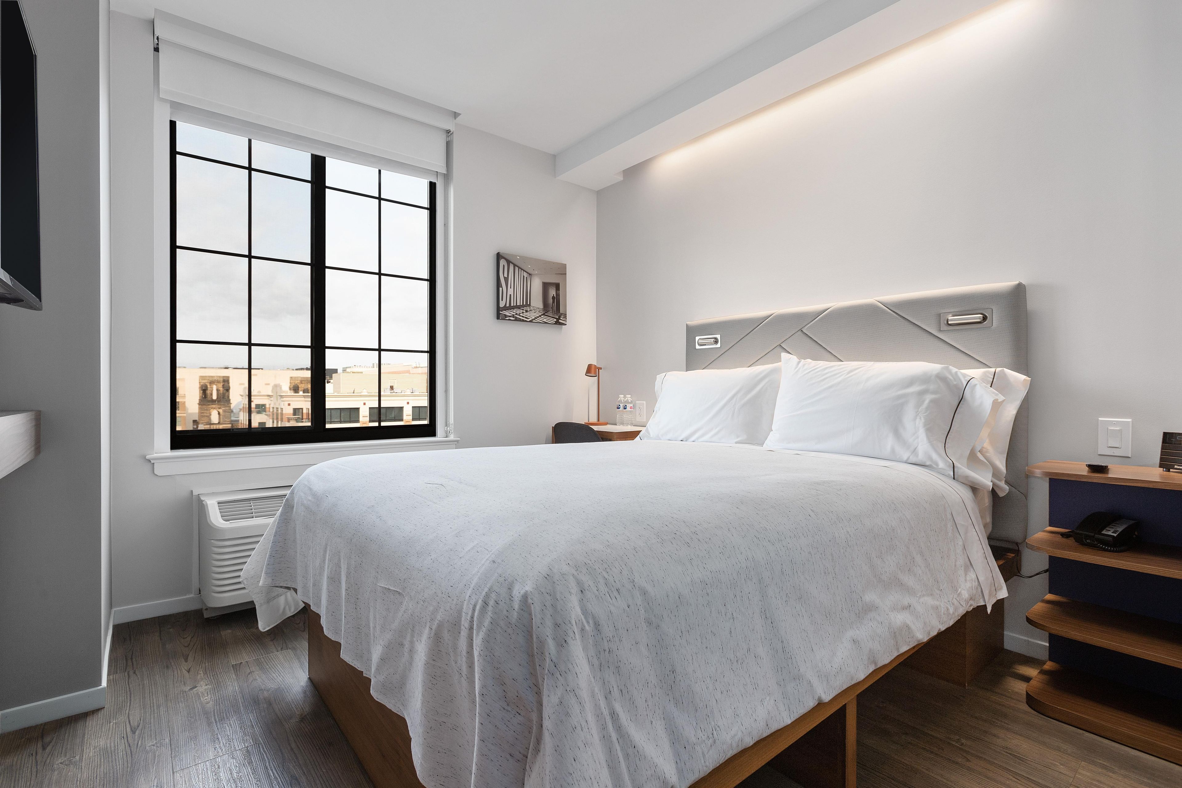 A modern hotel room with a large window, a bed with white linens, a bedside table with a lamp, and a city view.