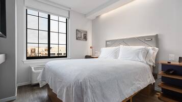 A modern hotel room with a large window, a bed with white linens, a bedside table with a lamp, and a city view.