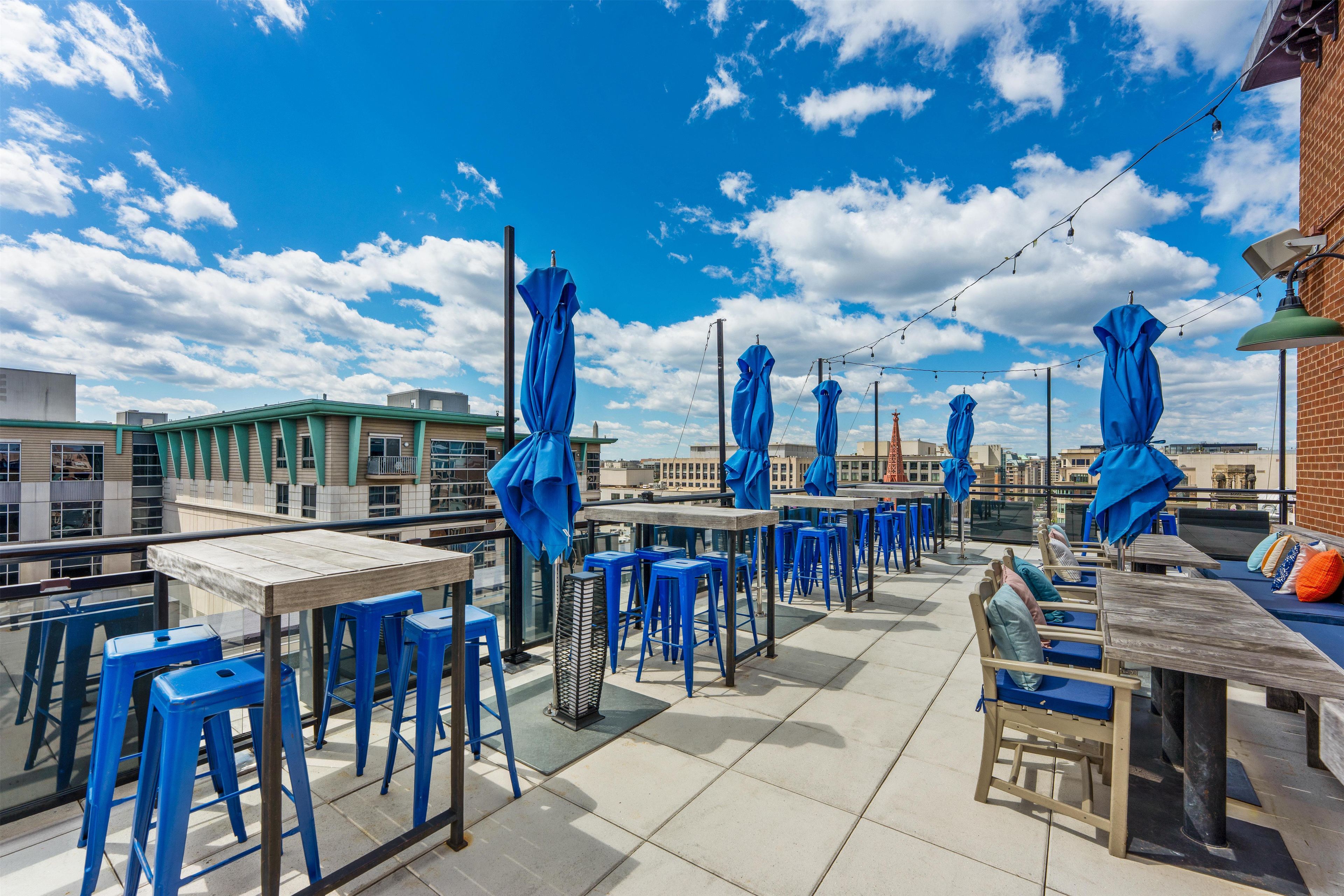 An outdoor terrace with tables and blue chairs, a brick wall, and a clear sky.