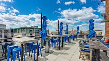 An outdoor terrace with tables and blue chairs, a brick wall, and a clear sky.