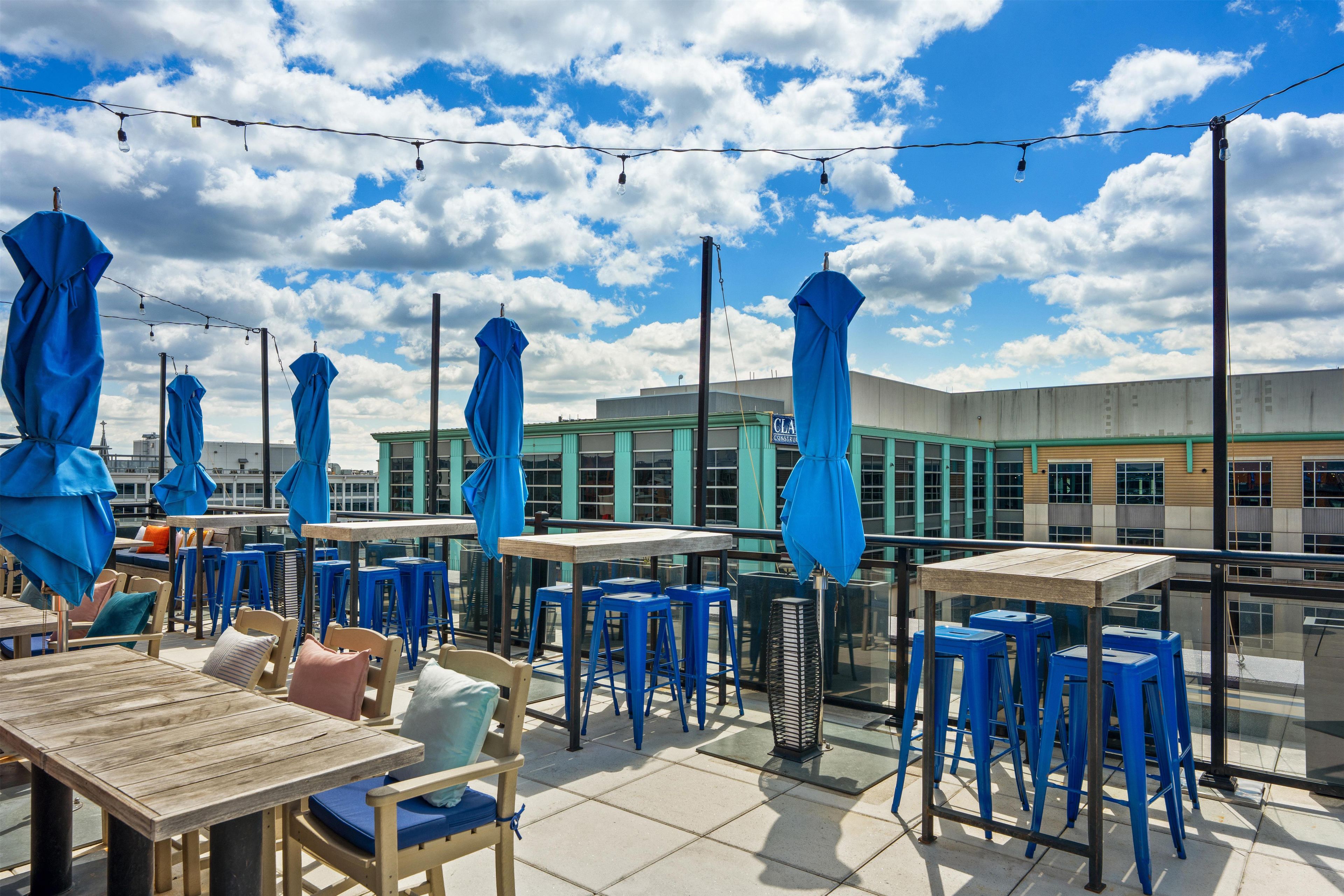 An outdoor seating area with tables and chairs, umbrellas, and string lights against a backdrop of a building and a clear sky.