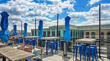 An outdoor seating area with tables and chairs, umbrellas, and string lights against a backdrop of a building and a clear sky.