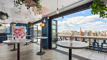 A rooftop terrace with round tables, hanging plants, and a city view.