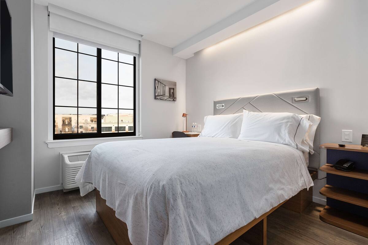 Ein modernes Schlafzimmer mit großem Fenster, einem Bett mit Kopfteil, einem Nachttisch und einem Beistelltisch.