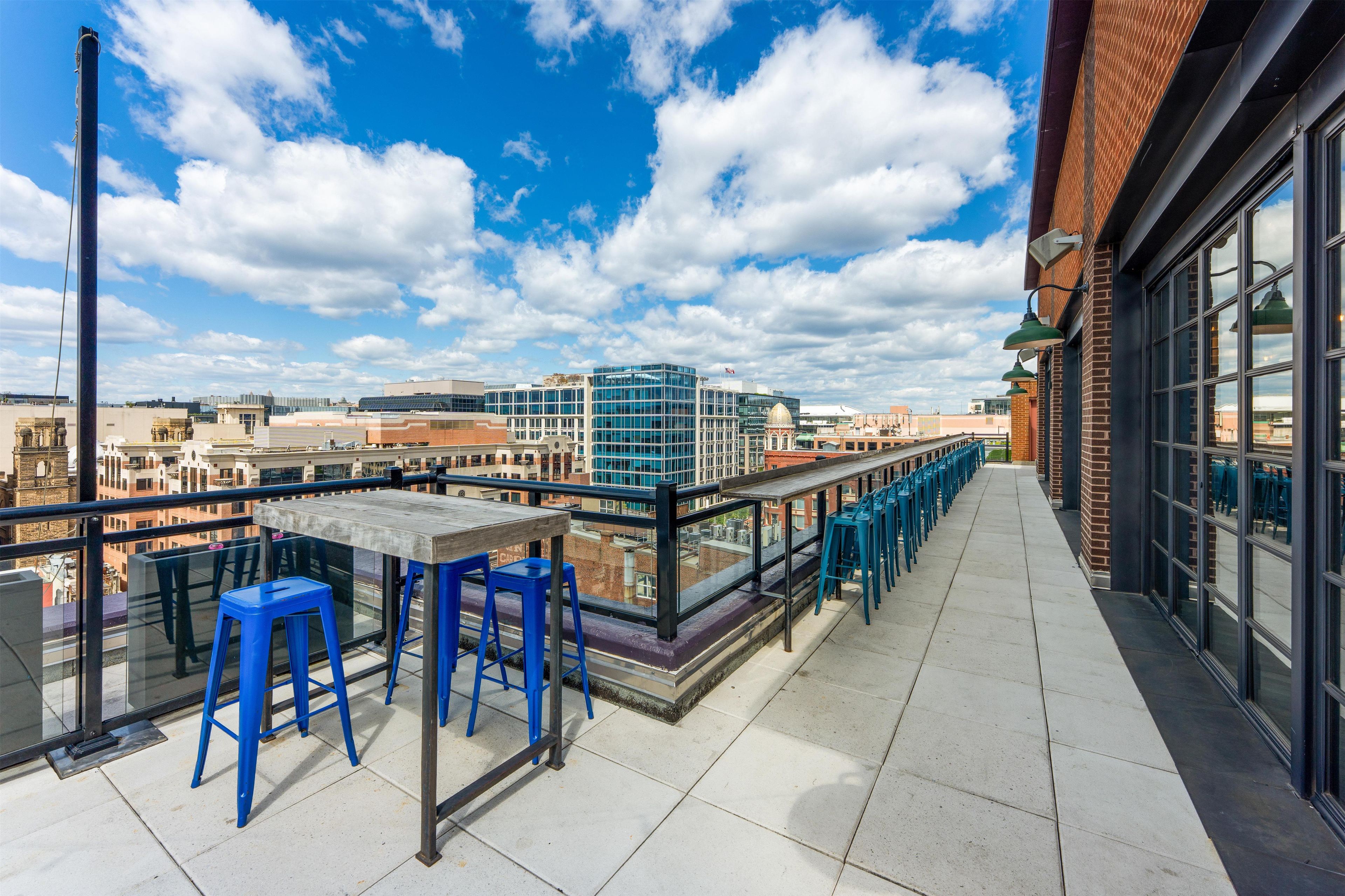 Une terrasse sur le toit, avec une table et des tabourets bleus, d’où l’on peut admirer un panorama sur la ville, avec ses bâtiments modernes.