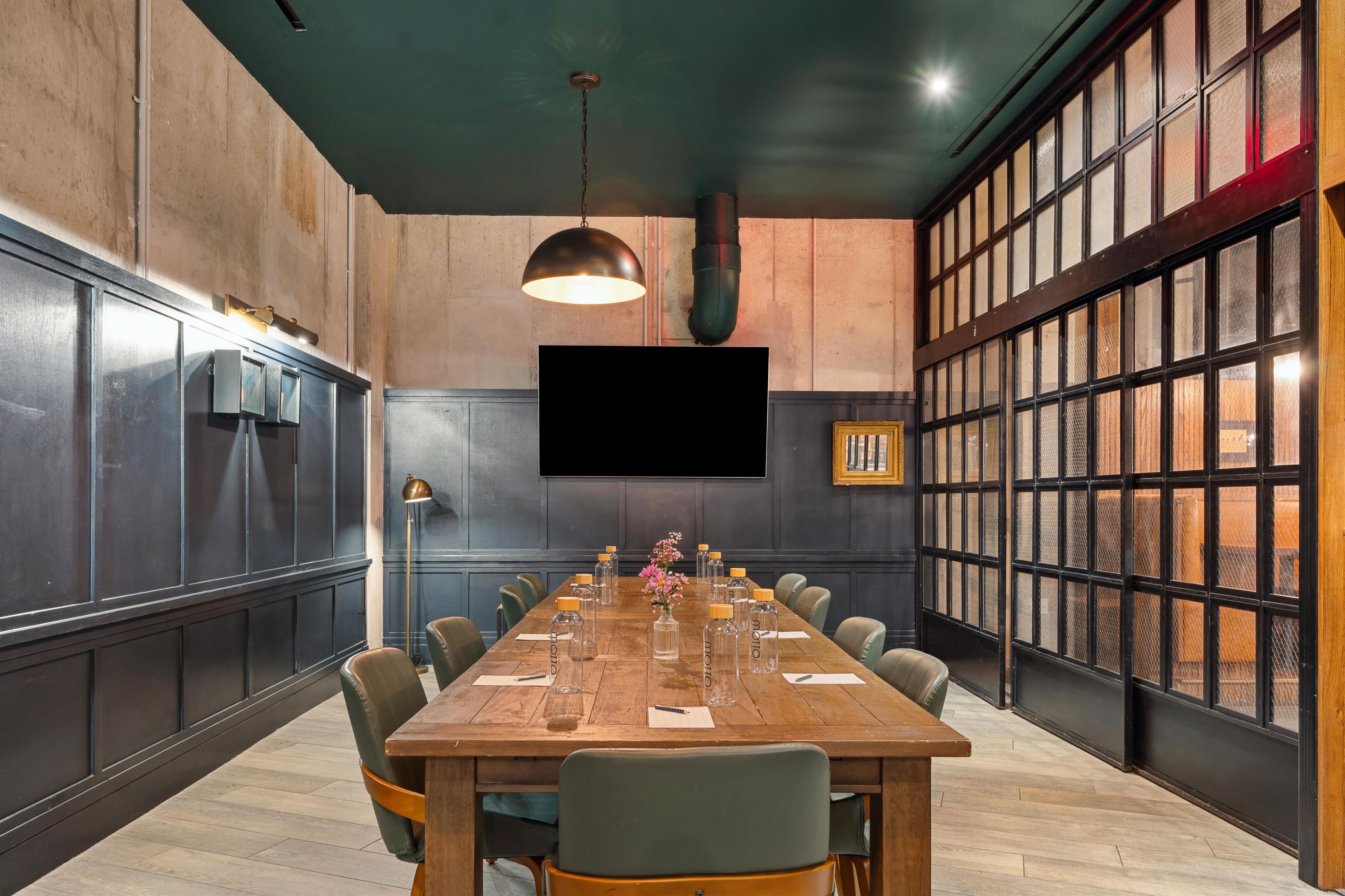 Un salón de conferencias con una larga mesa de madera, sillas, una pantalla de televisión y un amplio ventanal con persianas.