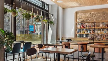 Een moderne bar met houten tafels en stoelen, diverse flessen op planken en hangende planten.