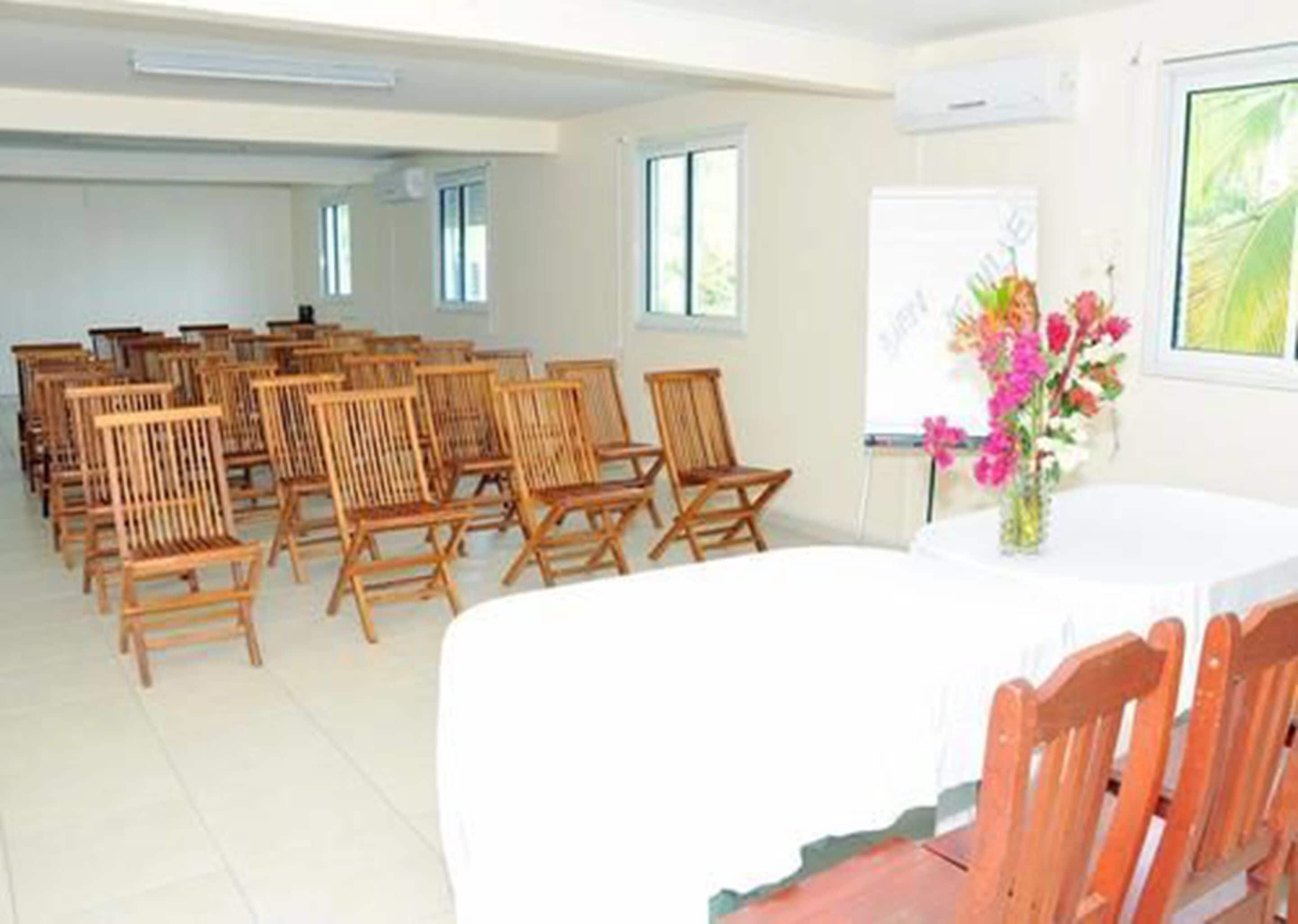 Uma sala de conferência com cadeiras de madeira dispostas em torno de uma mesa central, um quadro branco e uma janela com vista para vegetação.