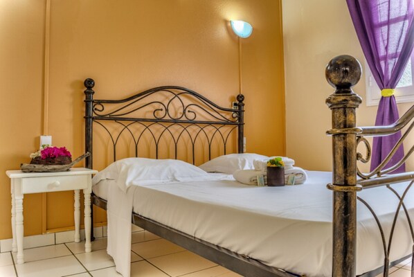 A neatly made bed with white linens, a bedside table with a basket of flowers, and a wall-mounted light fixture. - Le Paradis de l'Anse (Riviere-Pilote)