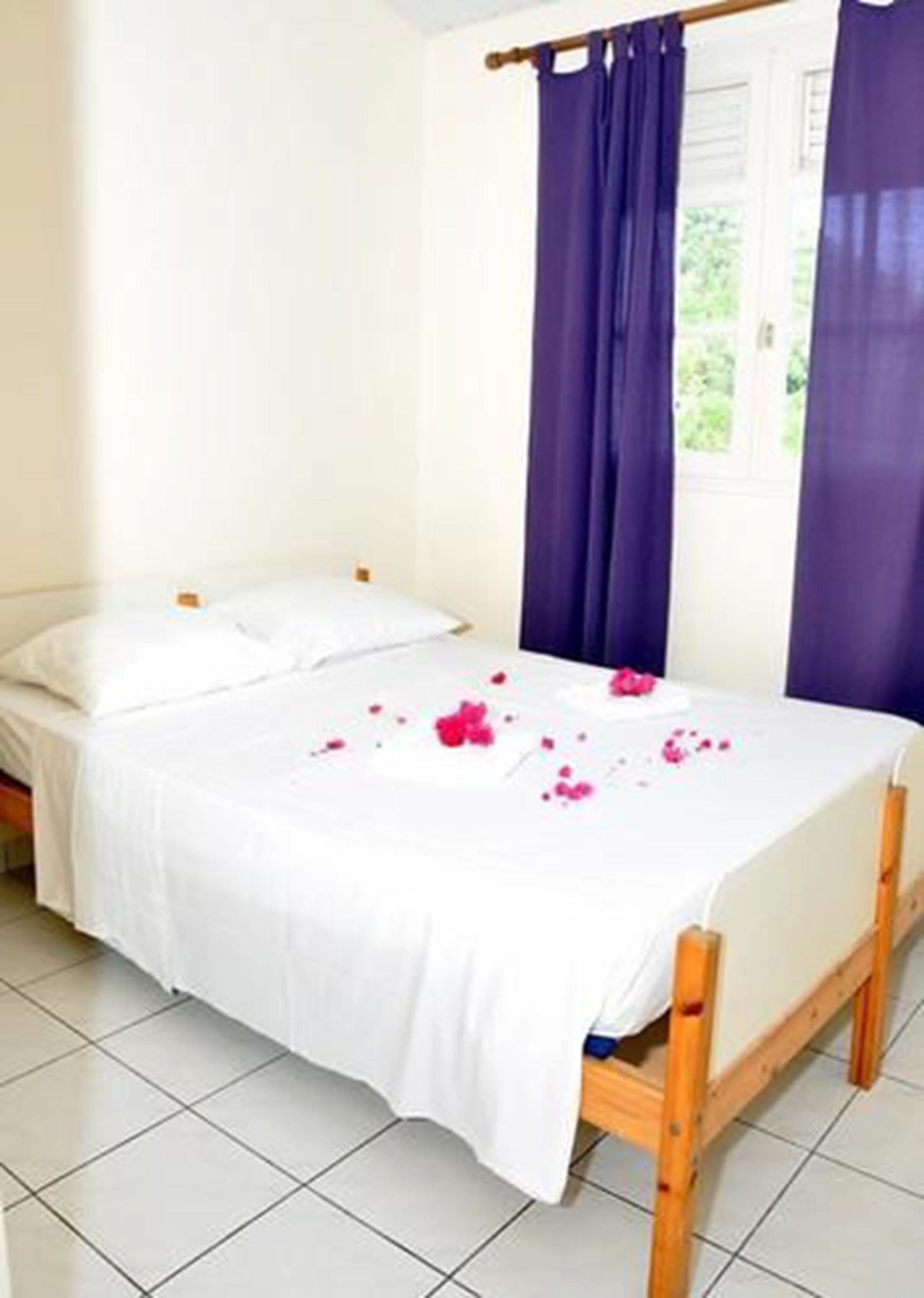 A single bed with white bedding and a wooden frame, a window with purple curtains, and a tiled floor.