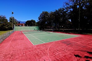 Una cancha de tenis al aire libre con superficie roja, rodeada de árboles y una montaña al fondo.
