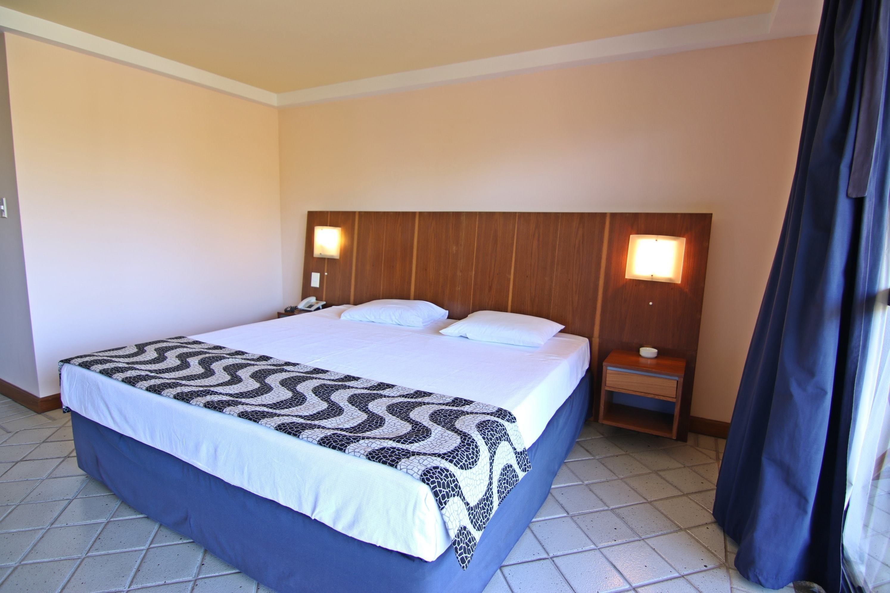 Quarto de hotel com uma cama grande, duas mesas de cabeceira e cabeceira com luzes fixadas na parede.