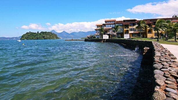 Un hotel costero con un muro de contención de piedra, vistas despejadas al mar y montañas al fondo.