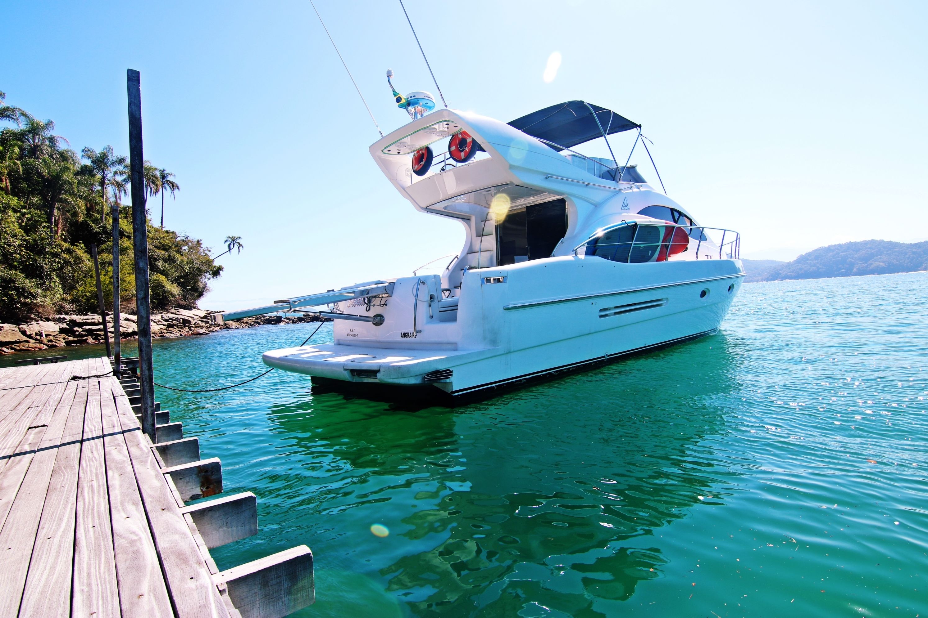 A white yacht docked at a wooden pier with a clear view of the water and distant hills.