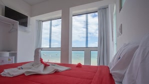 A hotel room with a large bed, a TV mounted on the wall, and a view of the sea through large windows. - Pousada Vela Mar (Maragogi)