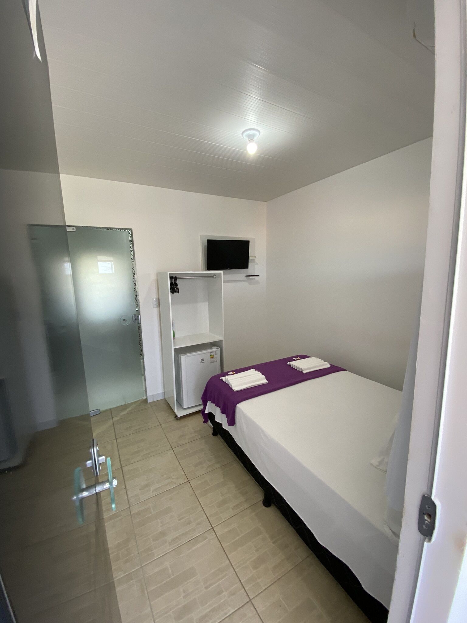 Habitación de hotel con una cama, un televisor montado en la pared, un refrigerador pequeño y un vidrio corredizo que da a otra habitación.