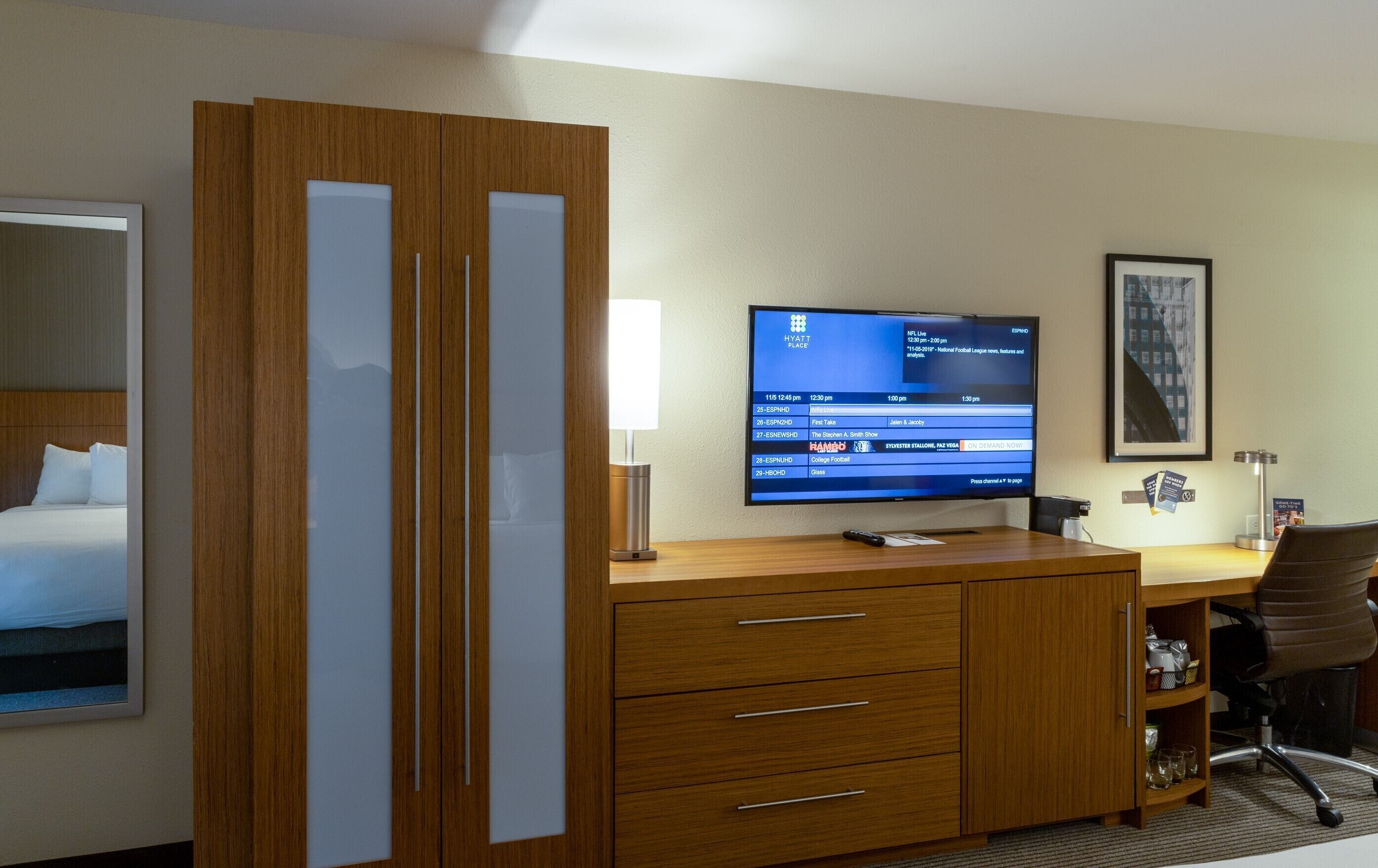 Habitación de hotel con un escritorio de madera, un televisor que muestra un canal deportivo, una cama y un cuadro enmarcado en la pared.