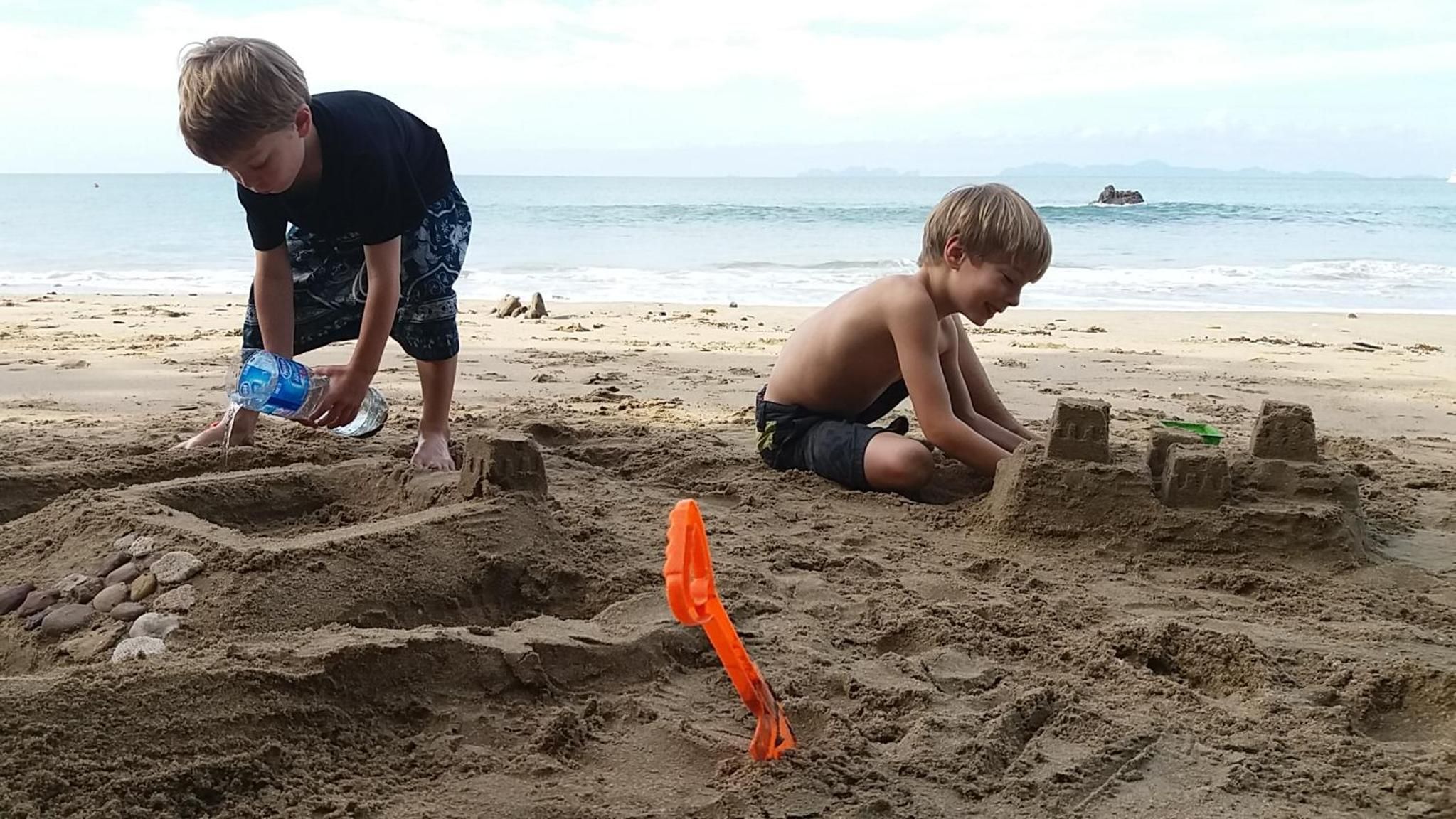 Zwei Kinder spielen an einem sandigen Strand, im Hintergrund Wasser und Felsen.