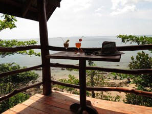 Terraza de madera con una mesa, dos bebidas y un sombrero con vistas a la playa.