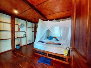 Una cama individual con mosquitero, suelo de madera, una mesita con un ventilador y un tapete azul en el piso.
