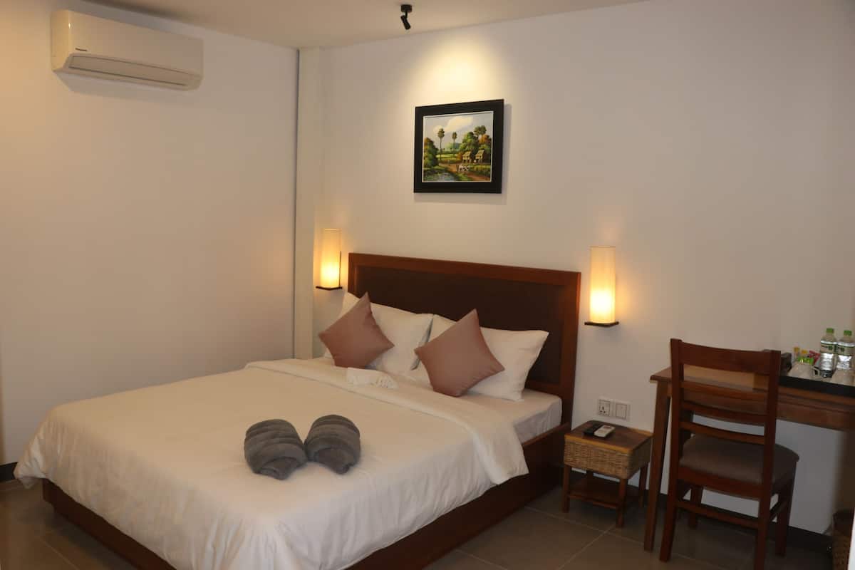 Cama bem arrumada com almofadas, mesa de cabeceira com abajur, cadeira e um quadro fixado na parede.