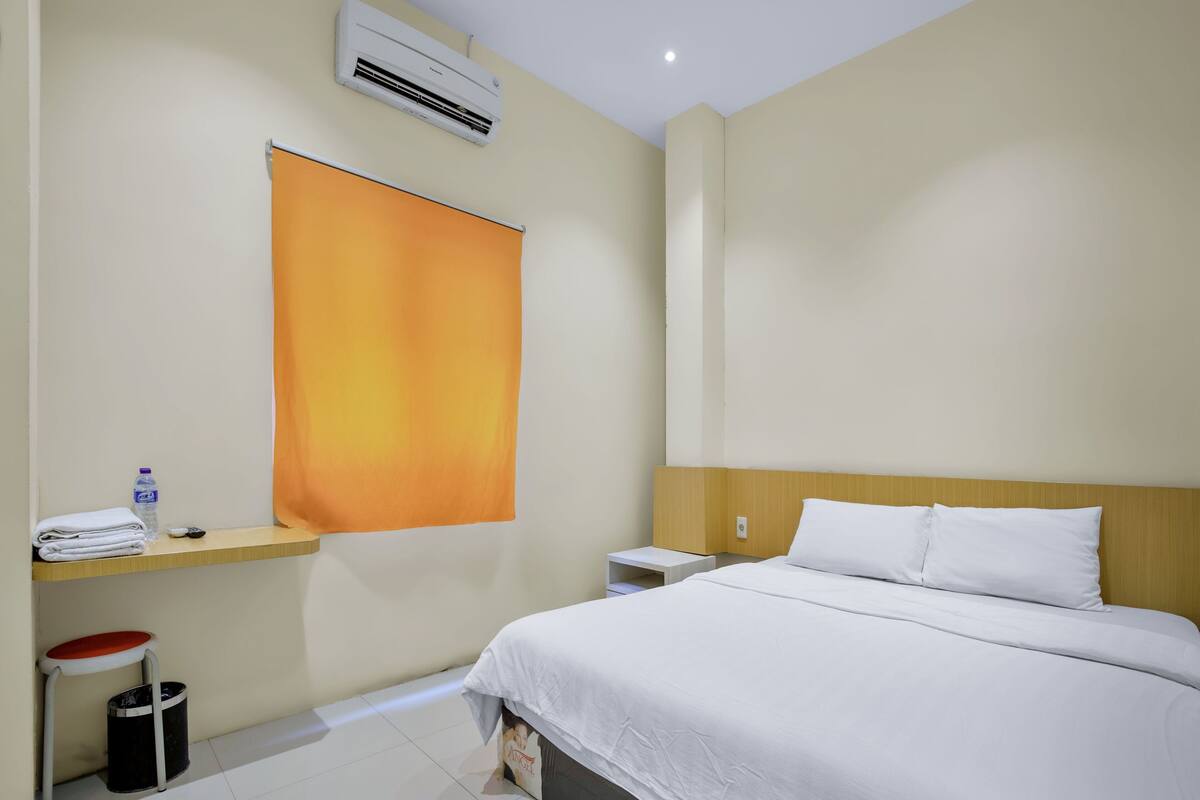 Habitación de hotel con cama, mesita de noche, una pequeña mesa con una botella y vasos, un cubo de basura y un amplio panel naranja que cubre la ventana.