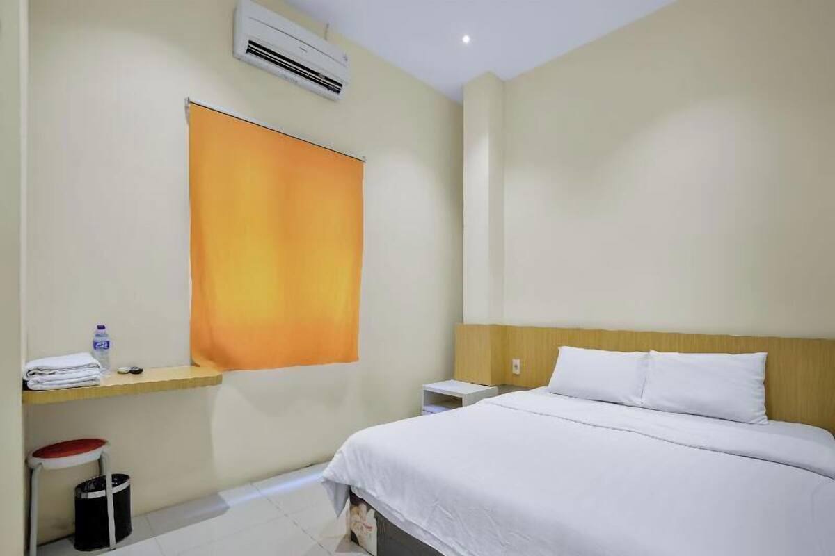 Una habitación de hotel con una cama, una mesita de noche, una pequeña mesa con una botella de agua, un cesto de basura y un aire acondicionado instalado en la pared.
