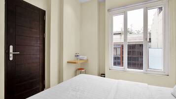 A bedroom with a dark wooden door, a white bed, a small wooden shelf with toiletries, and a window with a view of buildings outside.