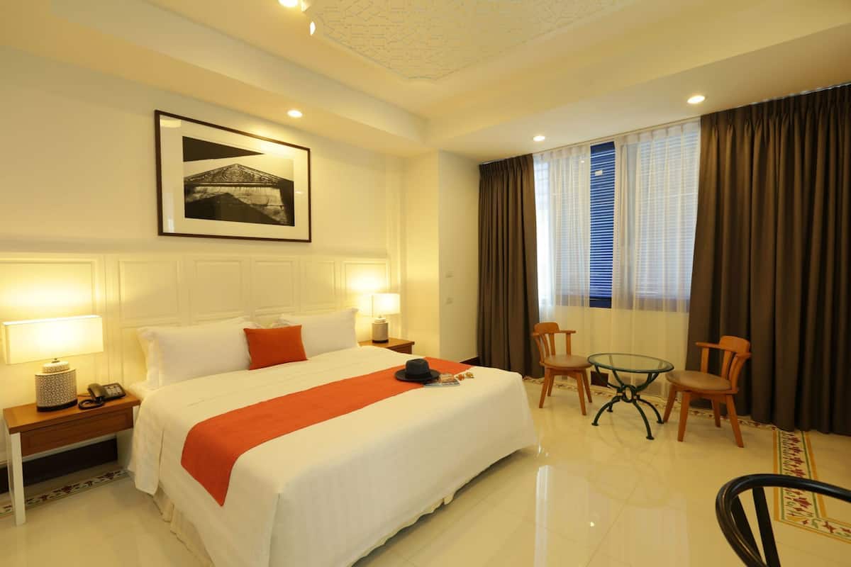 A hotel room with a large bed, two bedside lamps, a desk with a chair, a small table with two chairs, and a window with curtains.