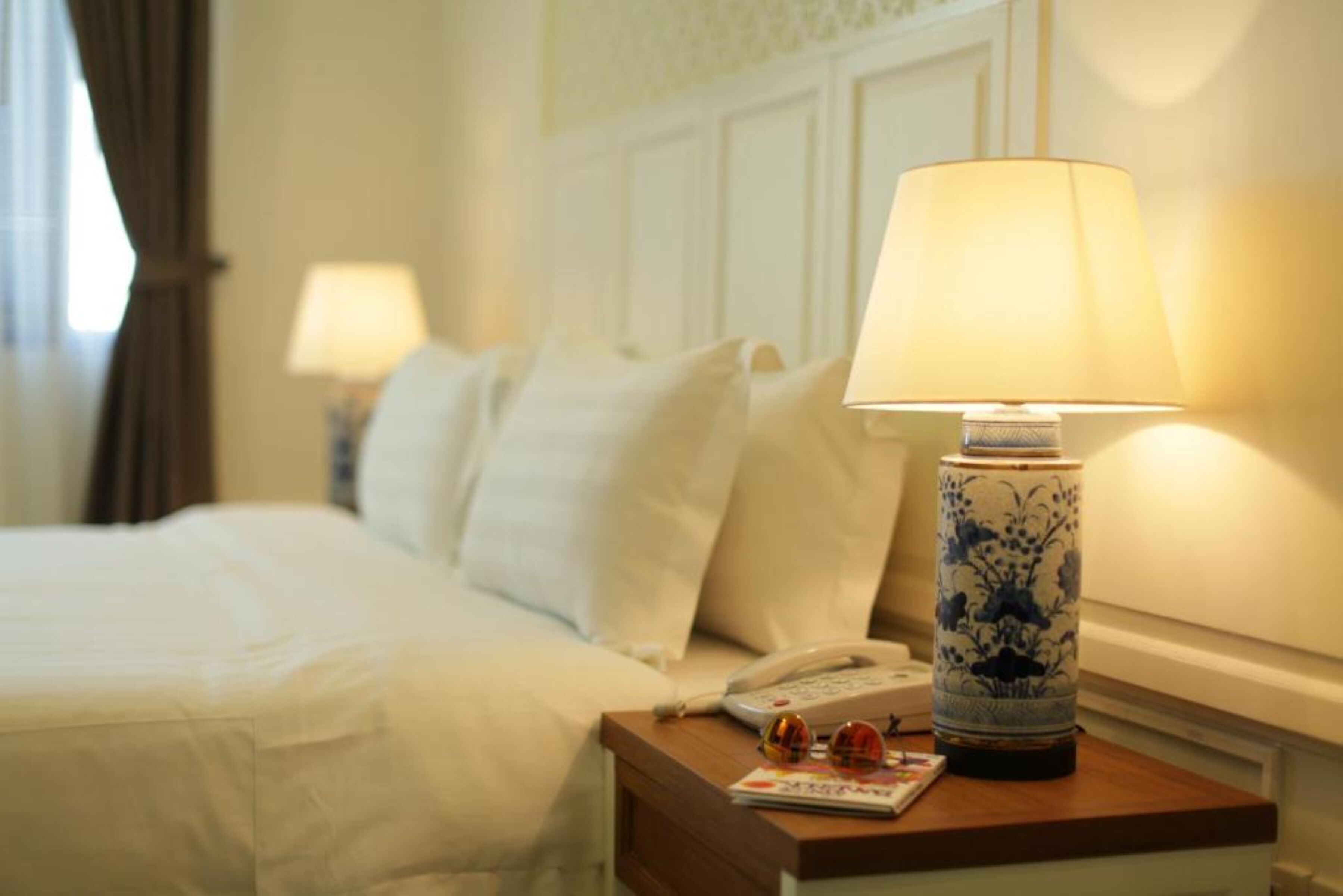 A hotel room with a neatly made bed, a bedside lamp, a telephone, and a magazine on the nightstand.