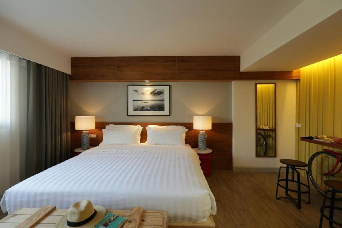 A hotel room with a large bed, two bedside lamps, a framed picture on the wall, a small table with a hat and book, and a bicycle in the corner.