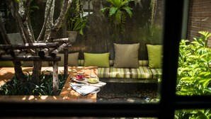 A view through a window of a room with a wooden table, a cushioned bench, and potted plants. - Nandha Hotel (Bangkok)