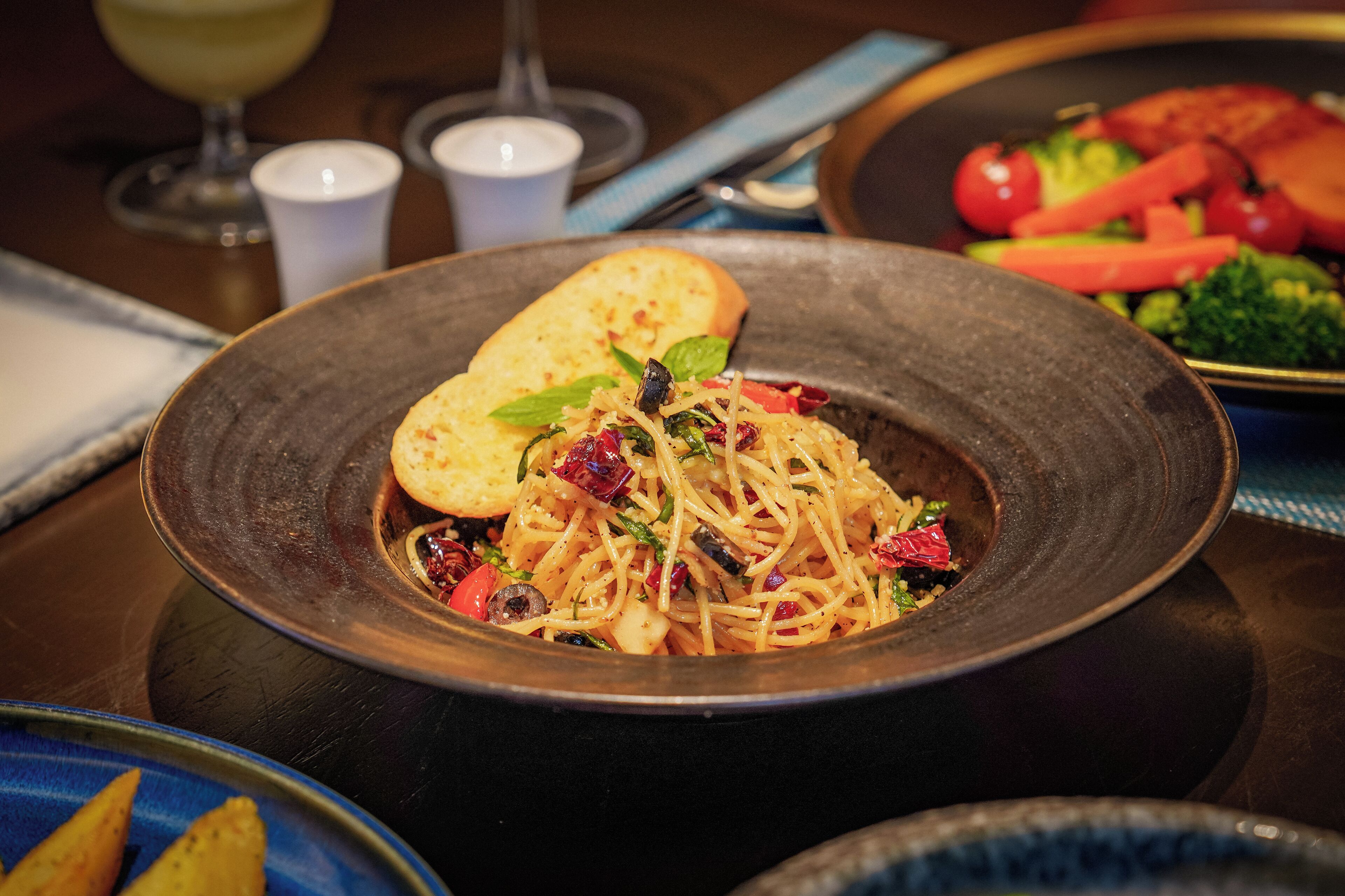 A plate of spaghetti with vegetables and a slice of bread on a dark plate.