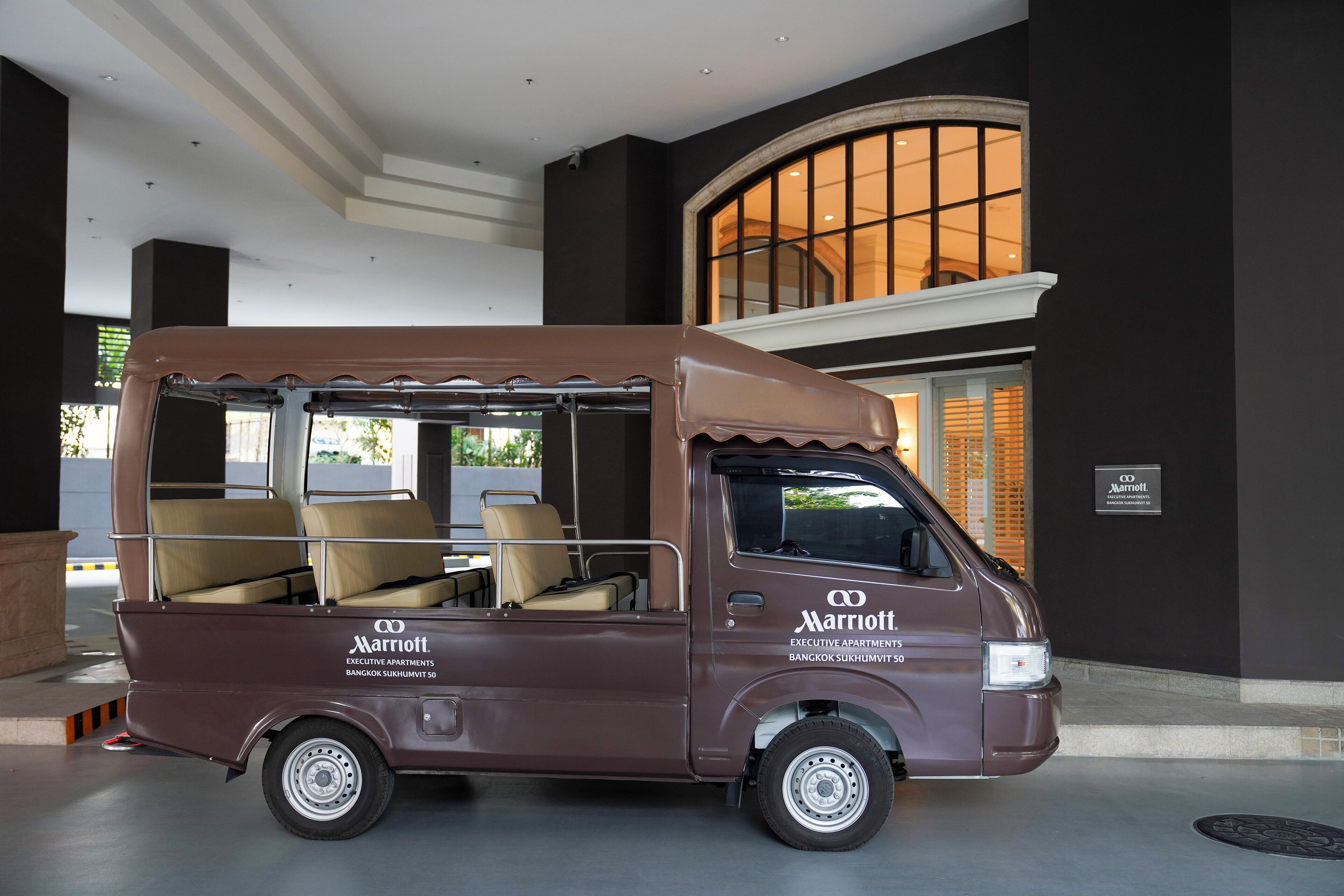 Een bruine bestelwagen met daarop 'Marriott Executive Apartments Bangkok Sukhumvit 50', geparkeerd voor een gebouw met een groot rondboogvenster.