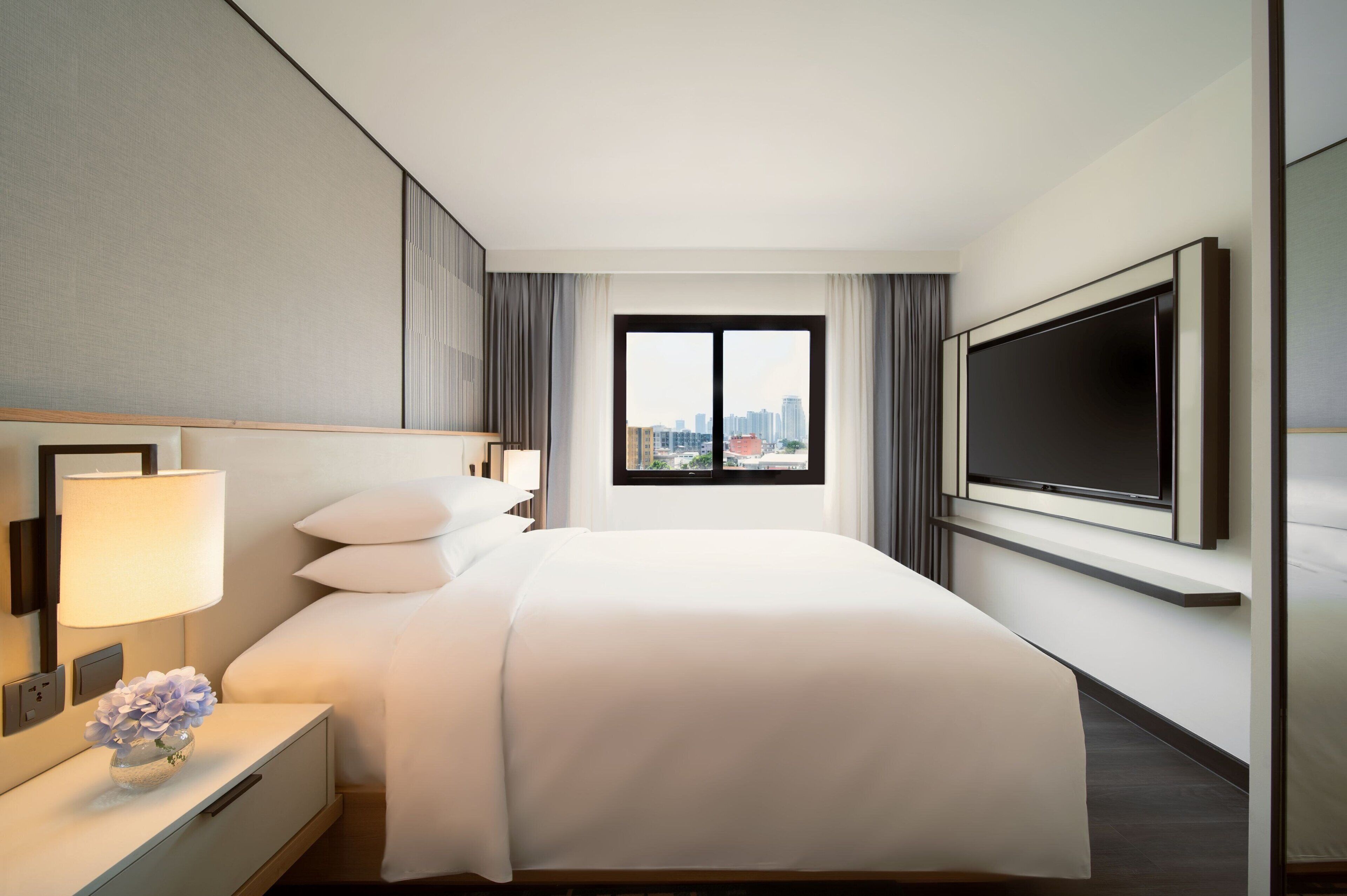 A hotel room with a large bed, a flat-screen TV mounted on the wall, a nightstand with a lamp, and a window offering a city view.