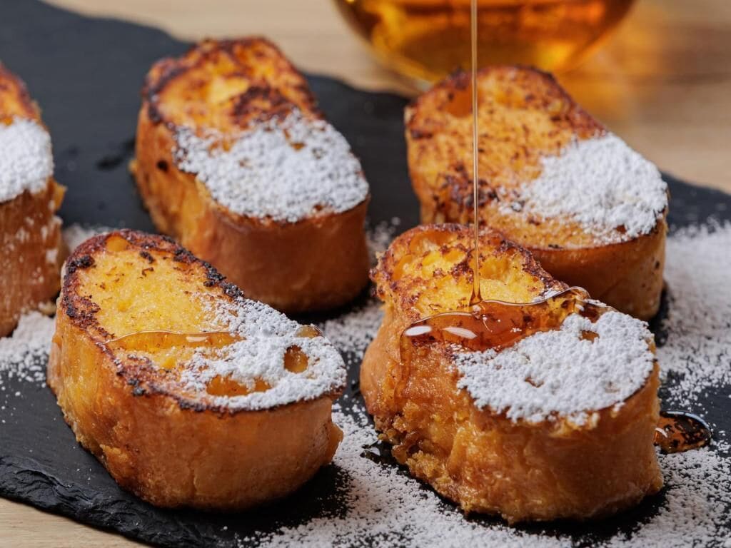 A close-up of breaded and fried items dusted with powdered sugar, with syrup being poured over them.