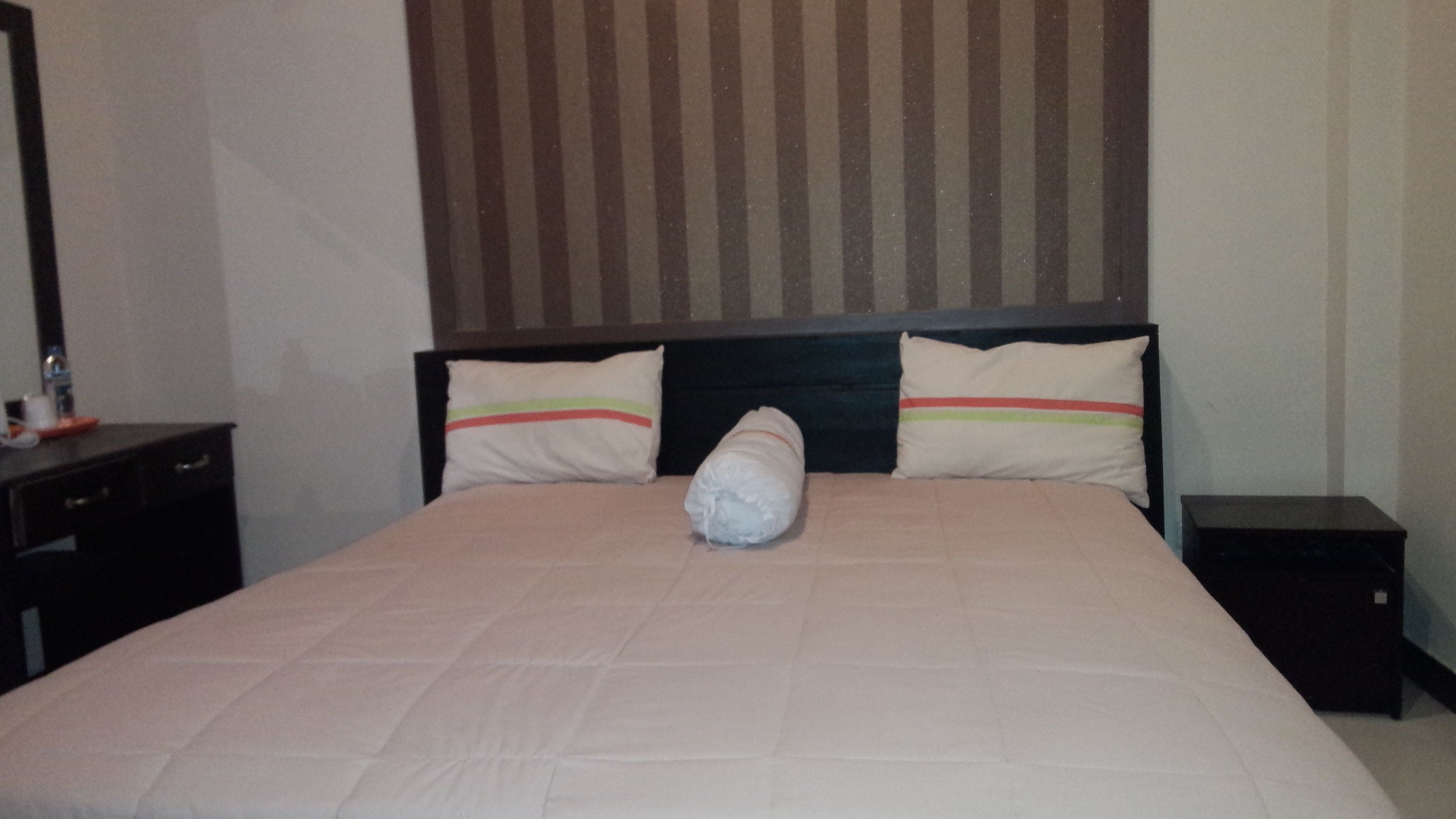 Ein ordentlich gemachtes Bett mit einem aufgerollten Handtuch, zwei gestreiften Kissen, einem dunklen Kopfteil und einem kleinen Nachttisch.