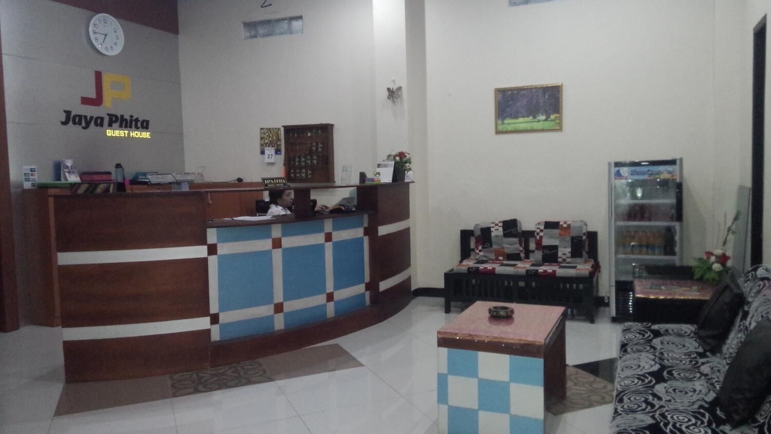 A reception area with a checkered counter, a clock, and a wall-mounted sign reading "Jaya Phita Guest House".