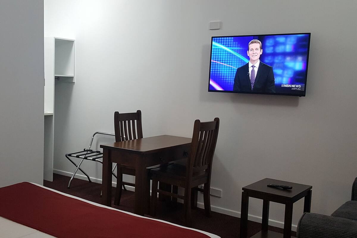 Une chambre d’hôtel avec un lit, un téléviseur fixé au mur, une table en bois, deux chaises et une petite table avec une télécommande.