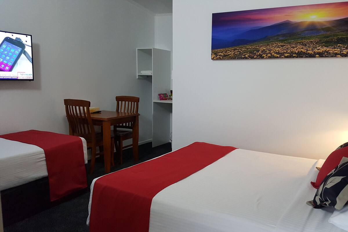 Une chambre d’hôtel avec deux lits simples, un bureau avec une chaise, une petite étagère et un tableau représentant un paysage au coucher du soleil.
