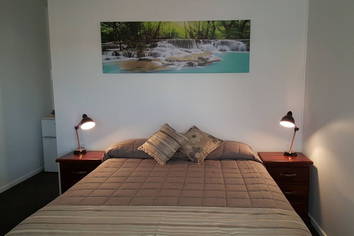 Une chambre à coucher avec un grand lit, deux tables de chevet avec des lampes, une petite table et un tableau représentant une cascade accroché au mur.