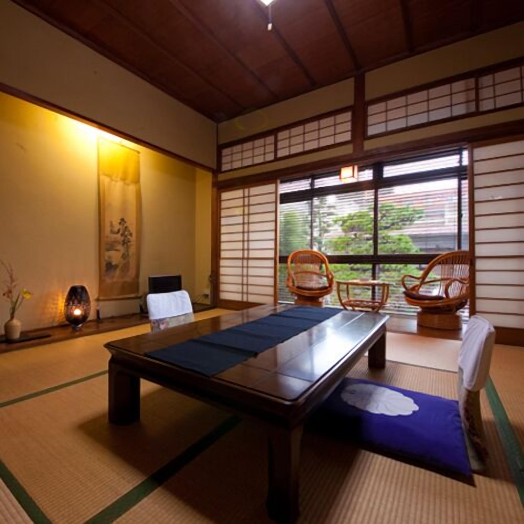 A tatami mat room with a low table, two chairs, a window with a view, and a traditional Japanese painting on the wall.