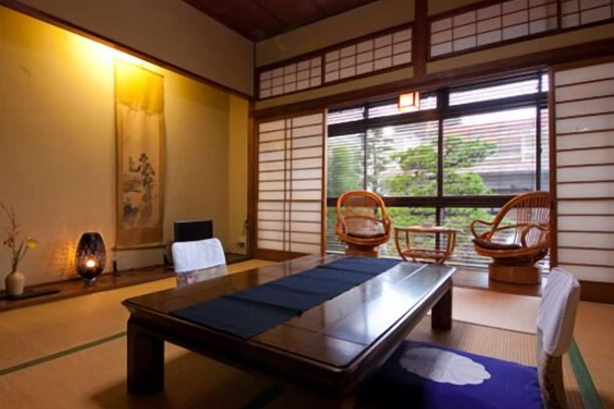 A tatami mat room with a low table, two chairs, a window with a view, and a traditional Japanese painting on the wall.