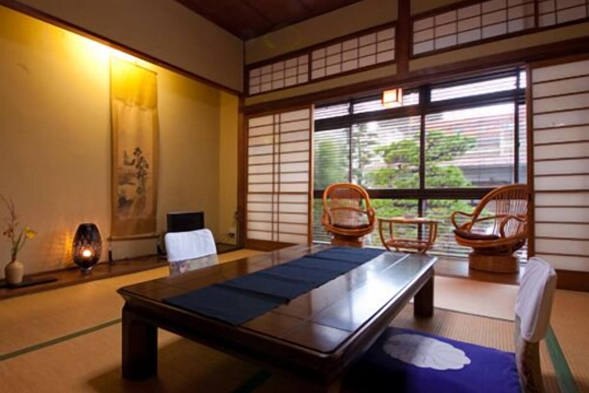 A tatami mat room with a low table, two chairs, a window with a view, and a traditional Japanese painting on the wall.