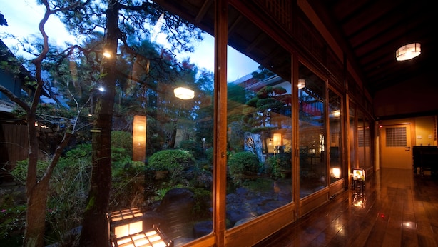 A traditional Japanese-style room with wooden flooring, large sliding glass doors, and a view of a garden with trees and a pond. - Ryokan Sennari (Beppu)