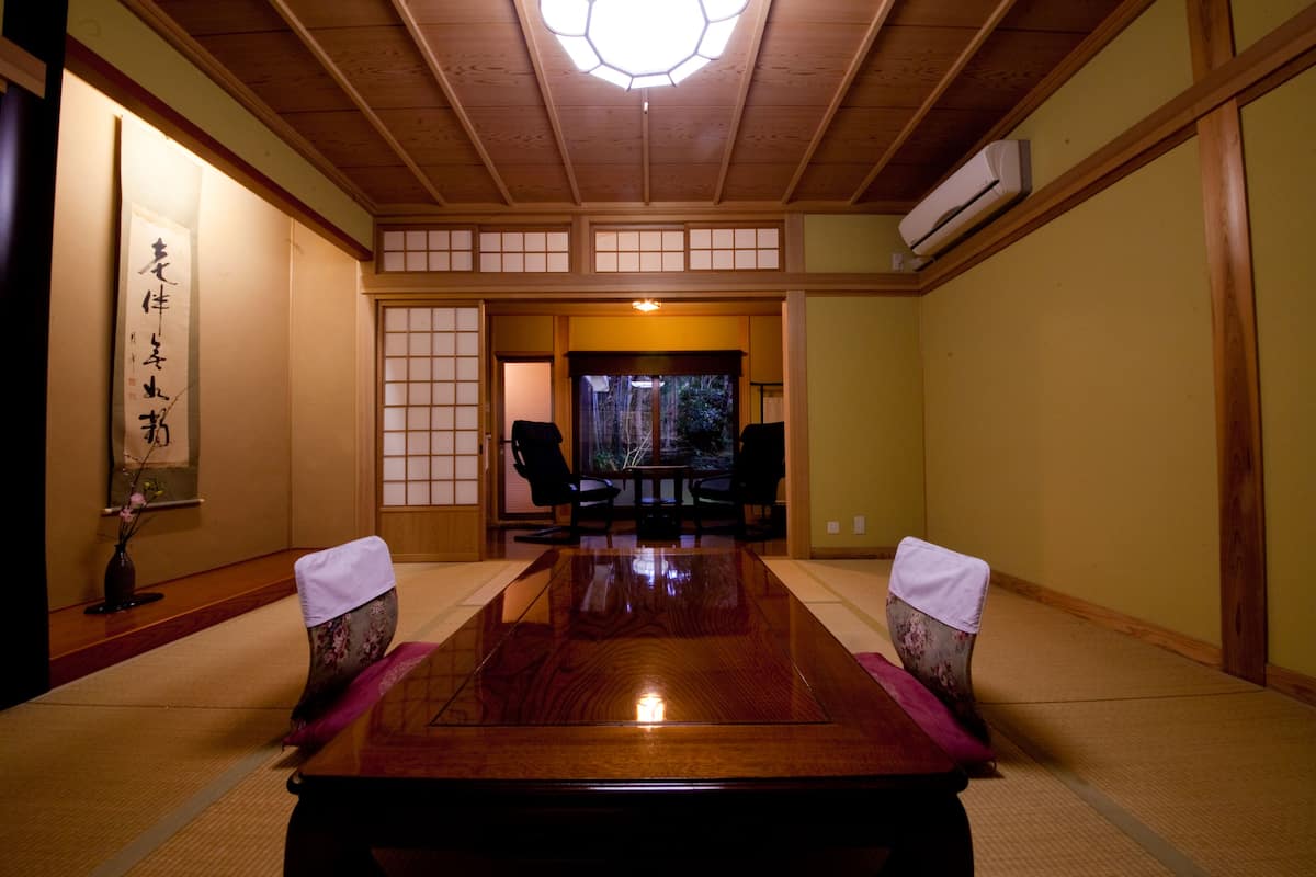 A traditional Japanese room with tatami flooring, a low table, two chairs, a large window, and a ceiling fan.