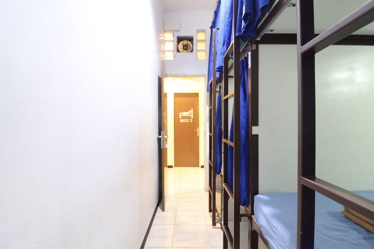 Un couloir étroit avec des lits superposés sur le côté droit, une porte marquée « CHAMBRE 3 » au fond, et un luminaire au plafond.